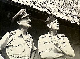 Half-length informal portrait of two men in short-sleeved tropical military uniforms with pilot's wings, standing in front of a hut and looking into the sky
