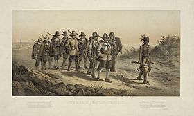 A group of nine 17th century militiamen carrying muskets and marching over a sandy path. A Native American man with feathers in his hair and carrying a musket is leading them. The soldier at the front of the group is wearing a helmet and a breastplate. In the background is a beach.