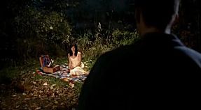 A woman is sitting on blanket with picnic materials around her. The scene is dimly lit, and a dark figure approaches.