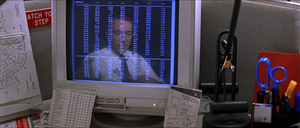A computer monitor on a busy cubicle desk. The monitor displays a spreadsheet in seven columns which span the height of the screen. The monitor also shows the reflection of a middle-aged man in a shirt and tie, sitting close to the desk and wearing a telephone headset. The contrasts—the monitor's dark background, and the lightness of the text and the man's shirt—make the reflection more prominent between and behind the numbers.