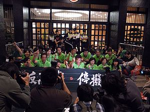 A group of protesters wearing green behind a black banner with Chinese writing in front of a building entrance surrounded by photographers