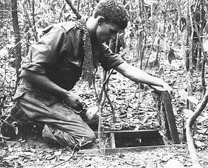 A soldier is kneeling over an open trapdoor on the jungle floor while holding a torch.