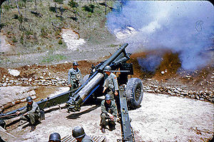 A colored photograph showing a howitzer recoils with fumes covering the front of the howitzer. A pair of GIs are standing besides the howitzer breech while another pair are crouching besides the howitzer trails. The heads of a third pair of GIs are at the bottom of the photograph near the howitzer shells.