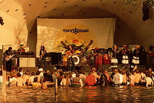 A crowd of people standing in water and listening to a band perform on stage.