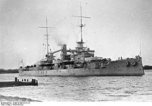 A large battleship lined with guns and equipped with two tall masts sits in harbor.