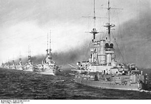 A long line of large, light gray warships sail through calm waters, each belching thick black smoke