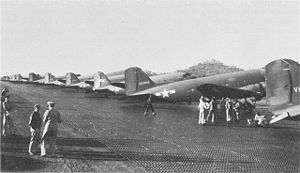 A steel plank runway with identical aircraft lined up along its length at right. Two small figures stand off to the left of the runway.