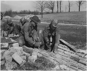 Five workmen. One is holding a shovel, while the other four are laying bricks to form a drainage ditch along the side of a road.