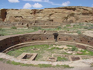Large circular depression outlined by a stone wall. The bottom is flat and grassy, and has a collection of rectangular stone foundations and smaller circles of stone. A great sandstone cliff towers in the background, and beneath the cliff are other stone foundations that are larger and higher.