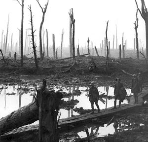 A group of soldiers walk across wooden duckboards that have been constructed over a waterlogged and muddy field. Shattered trees dot the landscape, with a low lying haze in the background