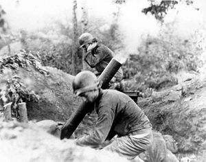 Two men in uniform brace as a mortar tube launches a rocket in the middle of a jungle