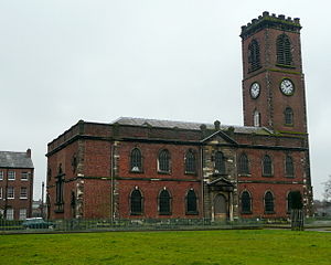 A two-storey brick church with stone dressings seen from the north; the tall slim tower has clock faces and a battlemented parapet