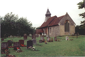 A small stone church with red tiled roofs seen through a churchyard from the southeast.  On its far gable is a bellcote with a pyramidal roof