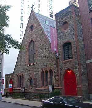 A brown stone triangular building with a square tower at the right on a city street. Its windows have pointed arches and there are similarly shaped red doors on either side. A large purple banner hanging from it has the word "Resurrection" on it
