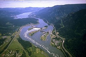 Aerial view of a large river winding through a mountainous gorge. It passes over a dam stretching in four segments from bank to bank across three intervening islands. Highways, passing by clusters of buildings here and there on both banks, run parallel to the river. Whitewater and foam curl downriver from one of the central segments.