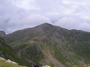 The side of a mountain covered with purple heather and green vegetation. The background is filled with blue-grey, puffy clouds.