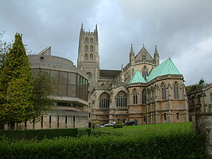 Ornate building with central tower. To the right is a stone building with green roof and to the left a new building with large glass windows.