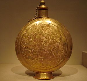 A circular, bulbous-disc-shaped golden canteen engraved with designs of a dragon and clouds, with a built-on stand and a cylindrical top that has a chain-link handle