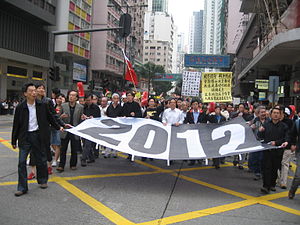 leaders of a protest march holding open a large banner with the figure '2012' in black-and-white print
