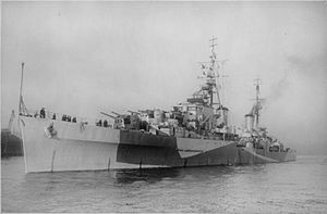 Black and white photo of a World War II-era warship. The ship is painted in camouflage and is armed with several gun turrets.