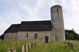 A stone church with thatched roofs seen from the north; on the left is a chancel with an apse, in the middle is the nave containing a Norman doorway, and on the right is a round tower