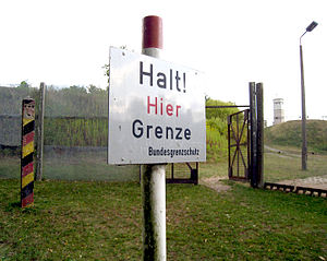 A white sign on a post with the German inscription "Halt! Hier Grenze" (Stop! Here border) and below, in smaller letters, "Bundesgrenzschutz" (Federal Border Guard). In the background a wire fence with an open gate, behind that are trees and a watchtower on the skyline.