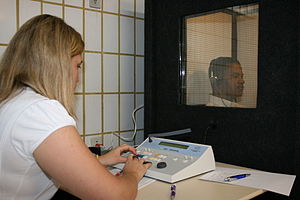 a female medical professional is seated in front of a special sound-proof booth with a glass window, controlling diagnostic test equipment. Inside the booth a middle aged man can be seen wearing headphones and is looking straight ahead of himself, not at the audiologist, and appears to be concentrating on hearing something