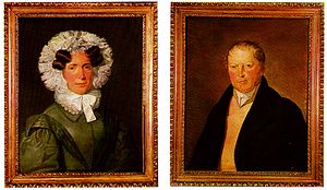 At left, a painted portrait of a woman in a black dress with a frilled hood and ruffled collar. At right, a painted picture of a man in a black coat wearing a cravat.
