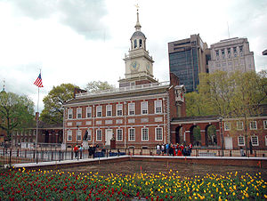 Front of Independence Hall on an overcast day. The steeple and facade of the building are visible, and an American flagpole stands on the building's right side. The foreground has a flower garden surrounded by a low brick wall with a fence on top. In the background, trees surround the building, and the Penn Mutual Tower and the Penn Mutual Life Building are visible, located to the right of the steeple relative to the viewer.