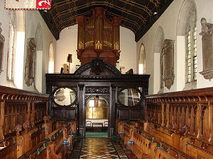 An organ with gilded pipes on top of a decorated wooden screen, with two rows of wooden benches on either side of an aisle