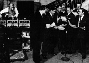 Seven suited men holding scripts and an eighth man operating a bank of turntables.