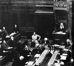 The speaker sits in a very ornate chair wearing a wig similar to that worn by judges. In front of him is a large table with books on it. Two people wearing wigs are seated at the head of the table and are writing. MacArthur is seated in a chair next to but not at the table. Men in suits are seated at the table. Behind them are rows of padded benches.