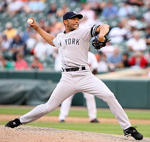 Mariano Rivera in a gray baseball uniform and navy blue cap stands on a dirt mound. He is striding forward to the right as he clutches a baseball behind his head. His uniform reads "New York" in navy blue letters across the chest. His face is contorted in concentration.
