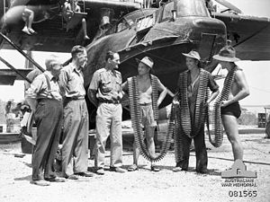 Members of No. 20 Squadron with lines of machine gun belts wrapped around them speak with members of the parliamentary war expenditure committee