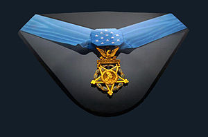 Gold medal, an inverted five-pointed star surmounted by an eagle, hanging from a formal blue ribbon in a display