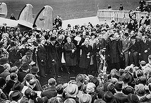 A large crowd on an airfield; British Prime Minister Neville Chamberlain presents an assurance from German Chancellor Adolf Hitler.