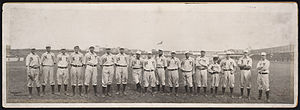 A single row of men in white baseball uniforms with high socks and white baseball caps standing on a baseball field; their uniforms read "NY" across the chest.