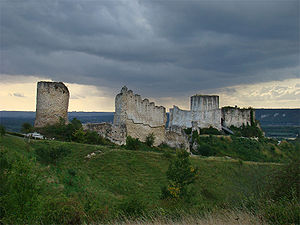 The ruins of a castle in grey limestone. It dominates the landscape. The castle's keep protrudes above the walls of the inner bailey; to the left a tower stands taller than the ruined walls.
