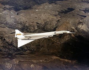 White delta-wing aircraft overflying mountains. At the front of aircraft are canards. Tips of wings are drooped down.
