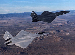 Gray and black jet fighters overflying rocky and barren terrain with the gray jet in the foreground.