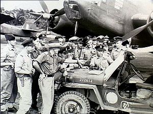 Approximately 15 men wearing military uniforms in discussion around a jeep, parked in front of a twin-engined aircraft
