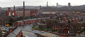 A view of a busy townscape, in which the land is urban and the scene is banal. The weather is overcast and the cloudy sky appears light-grey. In the foreground is a dark-grey tarmac dual-carriageway road, which sweeps up the middle of the photograph to its centrepoint. All around the road are two-storey red-brick houses. On the left-side of the midground are three large red-brick factories of around five-storeys high. On the horizon is a towerblock.