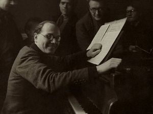A middle-aged, balding man with swept back dark hair, wearing a dark suit, seated at an upright piano. He faces the camera, his left hand on an open musical score, his right hand resting on the side of the piano. Four people can be seen in the background.