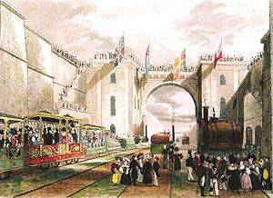 A large crowd standing in a deep railway cutting. On the railway tracks are three elaborately decorated carriages and a number of small locomotives.