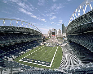 The interior of a stadium from the upper tier behind the south end zone during the day. The end zones and seating sections are colored blue. At the north end is a smaller seating area at the base of a tower. Several high-rise office buildings are in the distance.