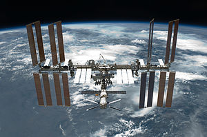A rearward view of the ISS backdropped by the limb of the Earth. In view are the station's four large, gold-coloured solar array wings, two on either side of the station, mounted to a central truss structure. Further along the truss are six large, white radiators, three next to each pair of arrays. In between the solar arrays and radiators is a cluster of pressurised modules arranged in an elongated T shape, also attached to the truss. A set of blue solar arrays are mounted to the module at the aft end of the cluster.