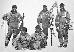  Five men in heavy clothing and headgear; three are standing and two seated on the ground. The standing men carry flags; all five have dejected expressions