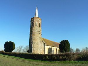 A flint church seen from the southwest, having a tall round tower with an octagonal bell stage and a lead spirelet.  The body of the church stretches to the right.