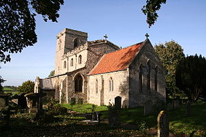 A stone church seeen from the southeast.  The chancel has a red tiled roof, the larger nave with clerestory has a battlemented parapet, and the tower has a plain parapet