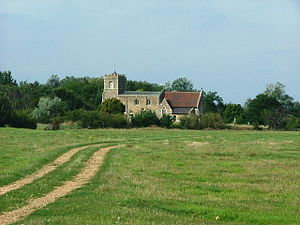 A field with a stone church in the distance, showing an embattled tower on the left, then the nave with a slate roof, and at a lower level the chancel with a red tiled roof